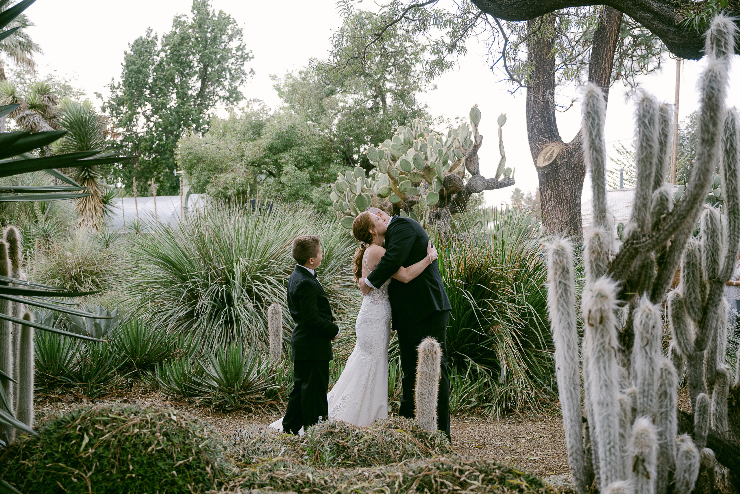 mom and dad hug during their first look on their wedding day surrounded by cactus with their son near by
