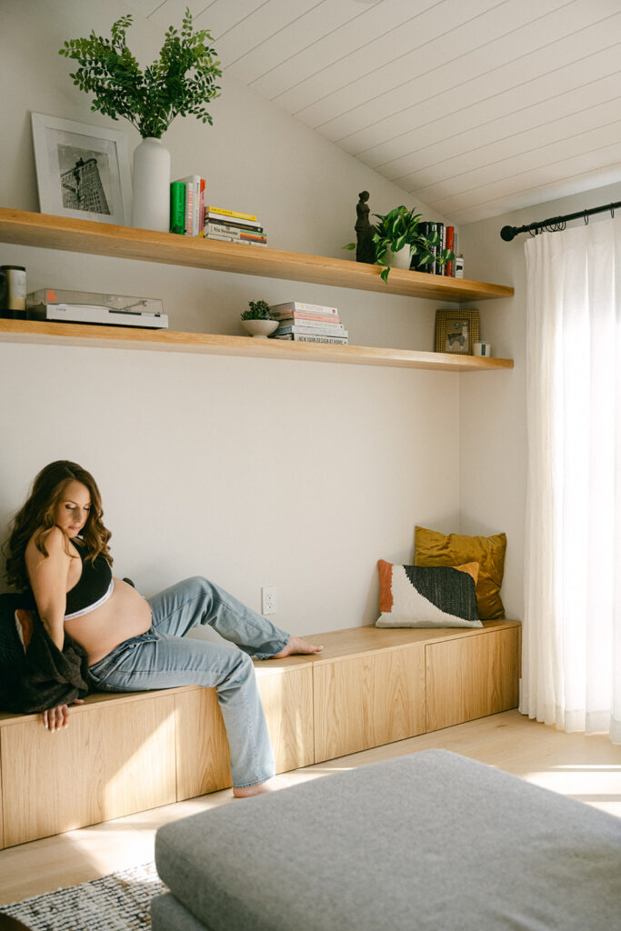 pregnant woman sitting on built in bench in sunlit room during in-home maternity photography session