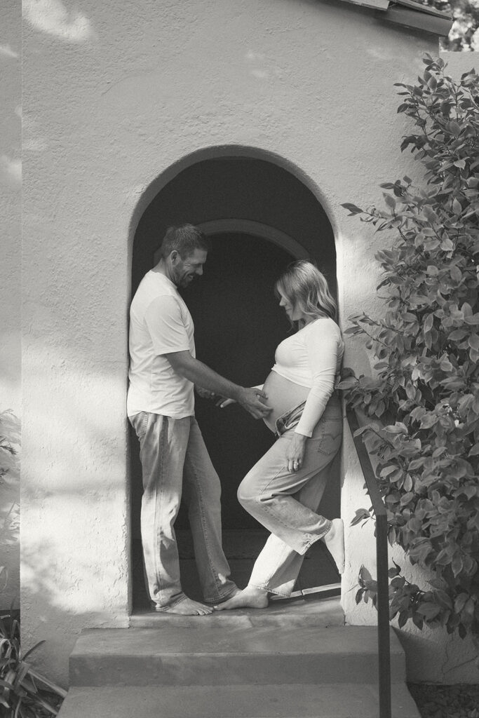 couple standing in doorway with partner touching pregnant belly during in-home maternity photography session