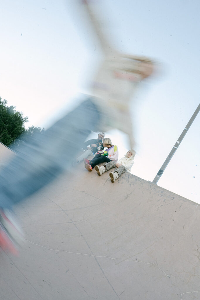family sitting on edge of skate ramp in St. Helena while child moves quickly in foreground during candid outdoor session