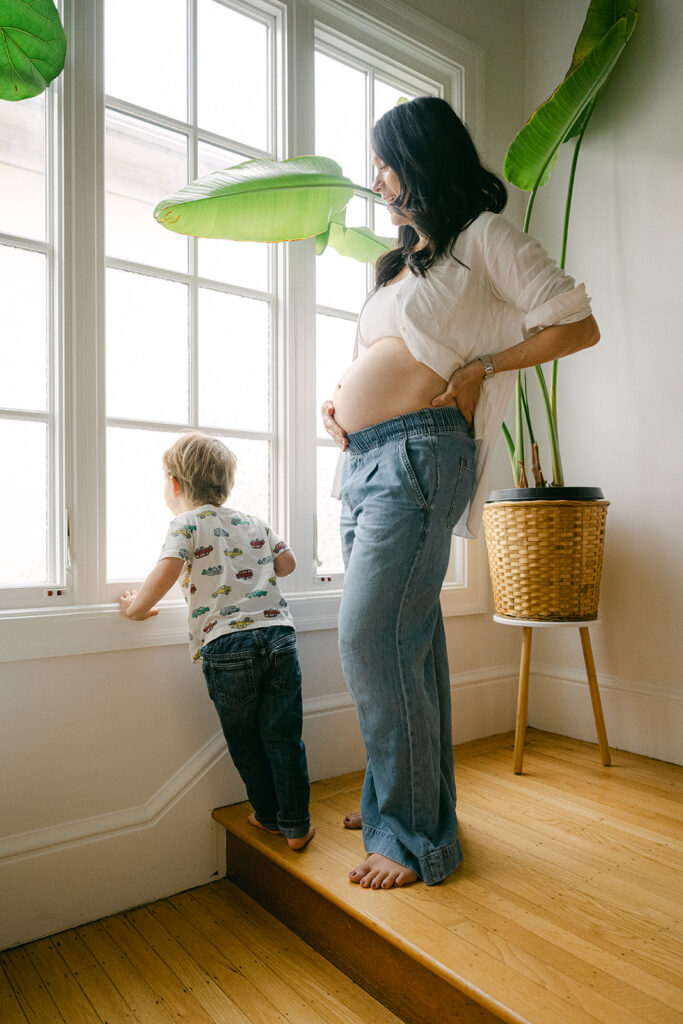 pregnant mother standing by window with toddler during in-home maternity photography session