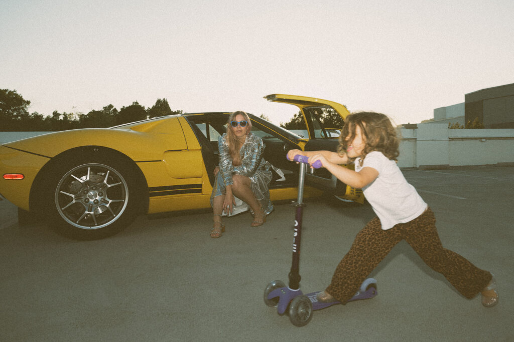 child riding scooter in foreground while mother sits in yellow sports car during candid outdoor family session at sunset