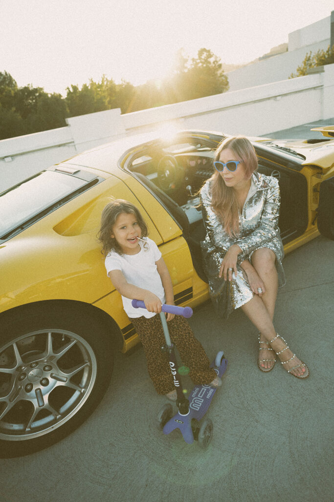 mother sitting in yellow sports car with child on scooter during playful lifestyle family photography session at sunset