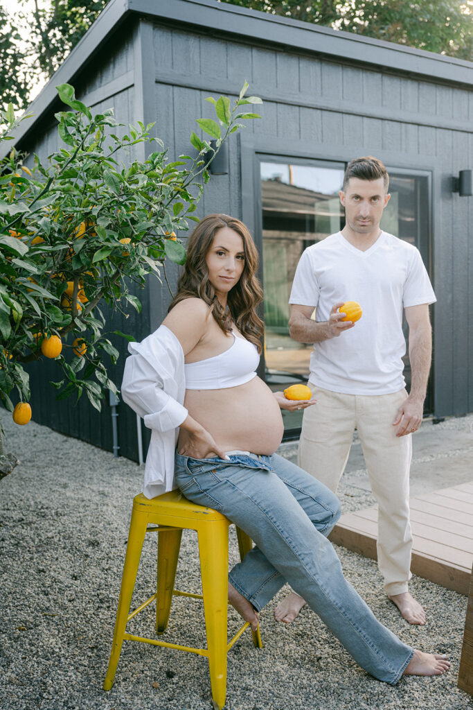 artistic maternity portrait of expectant mother seated on stool near lemon tree with partner holding fruit outside modern home