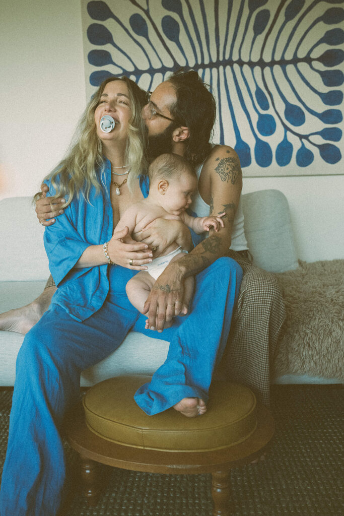 in-home family photography session in Benicia with mother holding baby and partner kissing her cheek on couch