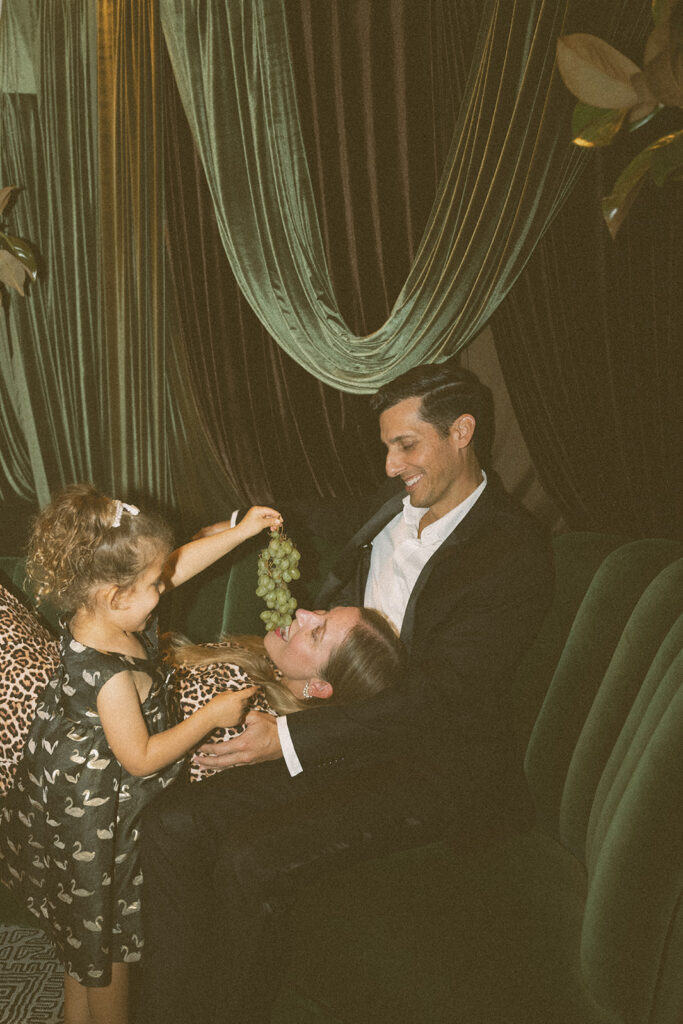 child holding grapes over parent while family sits together on couch during playful lifestyle family session